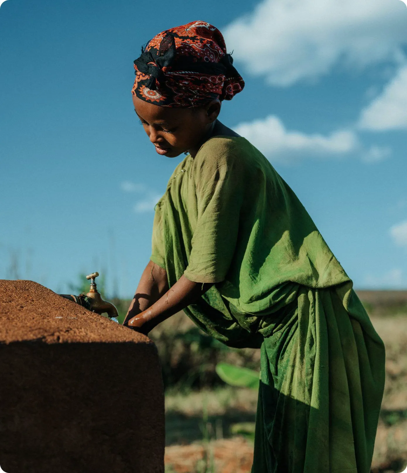 A young girl in a green dress and colorful headscarf washes her hands at an outdoor water tap under a clear blue sky.