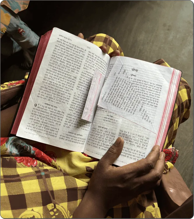 A person wearing a yellow checkered garment holds an open Hindi book in their lap, with a paper slip marking one of the pages. The setting appears indoors with soft natural light.