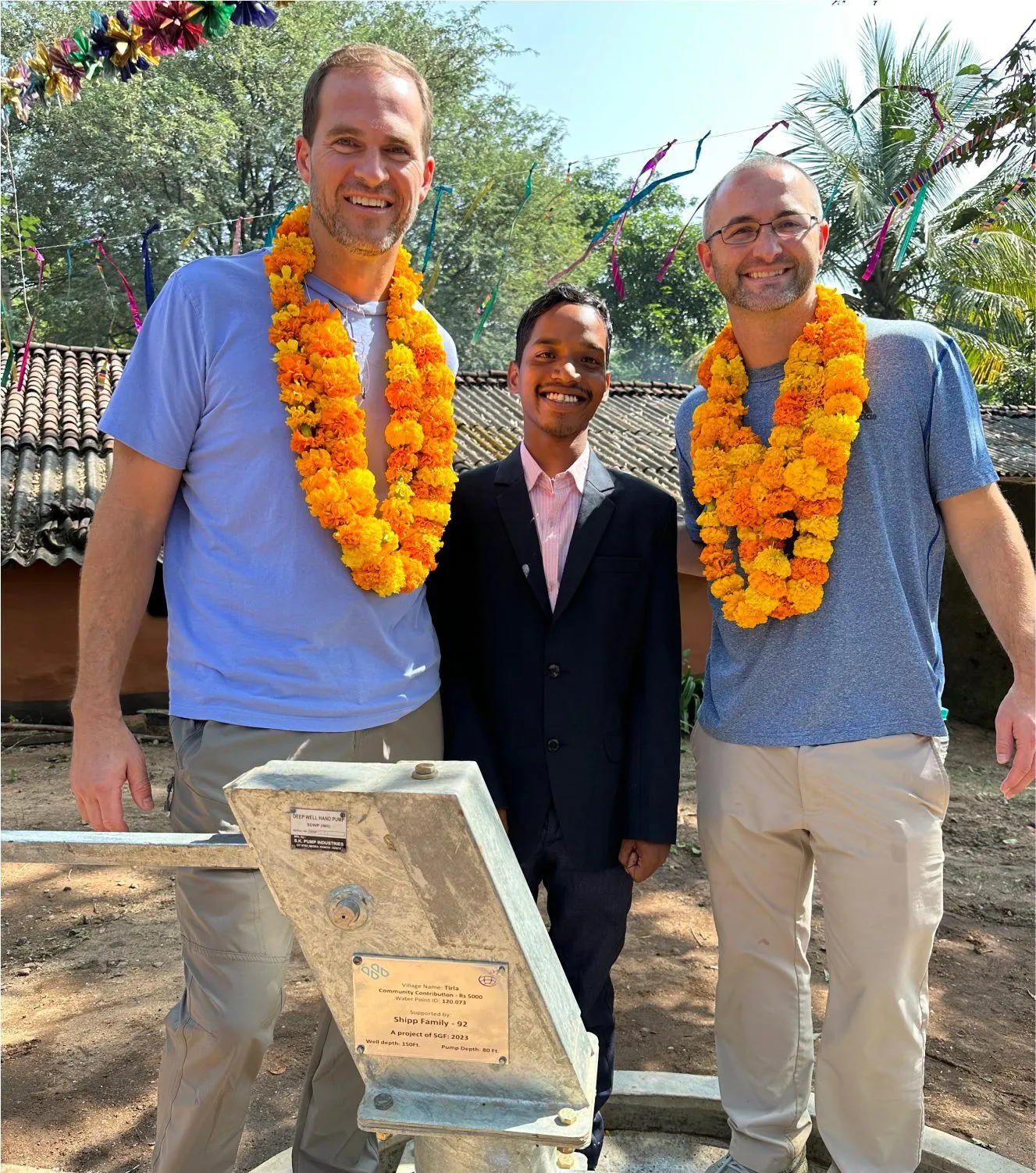 Two men wearing orange marigold garlands stand beside a smiling boy in a suit next to a metal water pump, with trees and colorful decorations in the background.