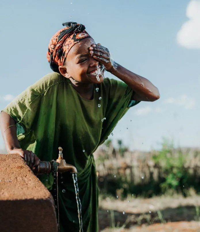A smiling child in a green shirt splashes water on their face from an outdoor tap, with water droplets visible in the air and greenery in the background under a clear sky.
