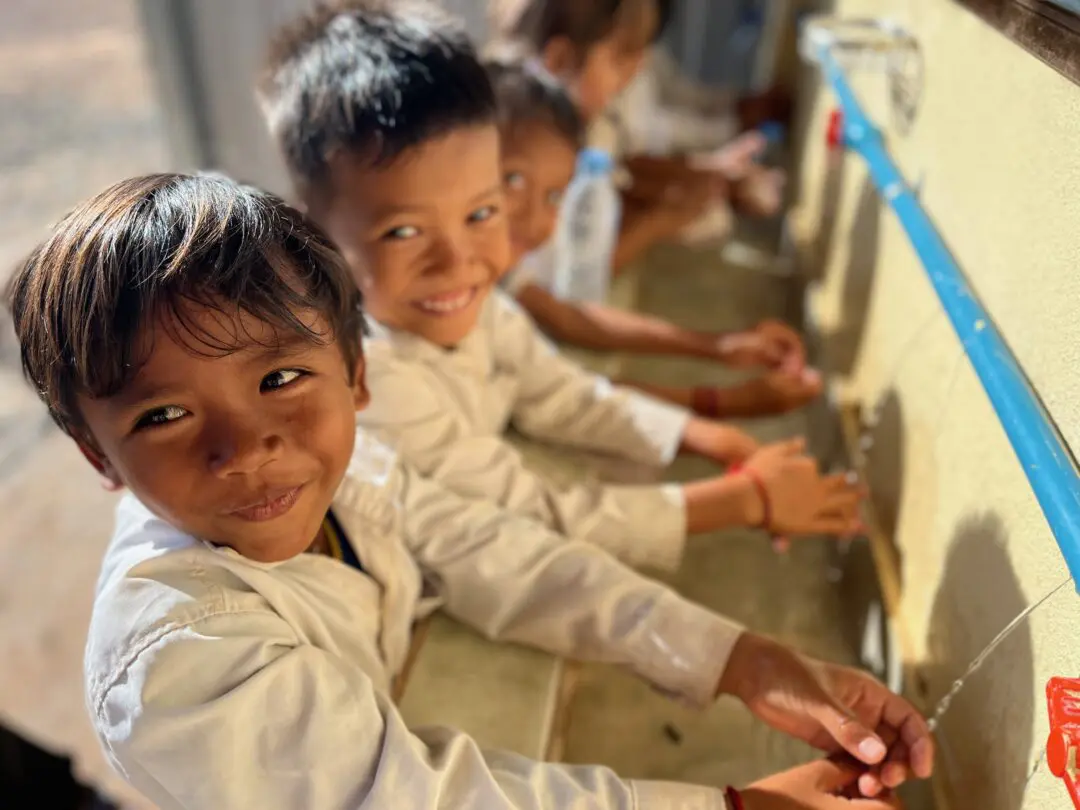 Two young children in white shirts smile at the camera while washing their hands at a sink, with other children washing their hands in the background.
