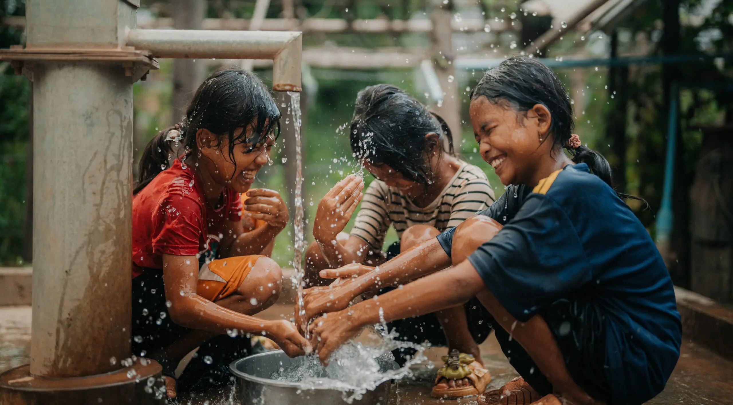 Three smiling children crouch around a water pump, playing and splashing water together, with droplets flying in the air. They look joyful and are washing their hands and faces outdoors.