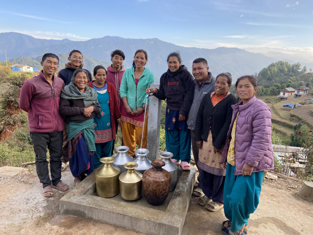 A group of people smiles and stands around several large water containers at an outdoor water station, with mountains and a rural landscape in the background.