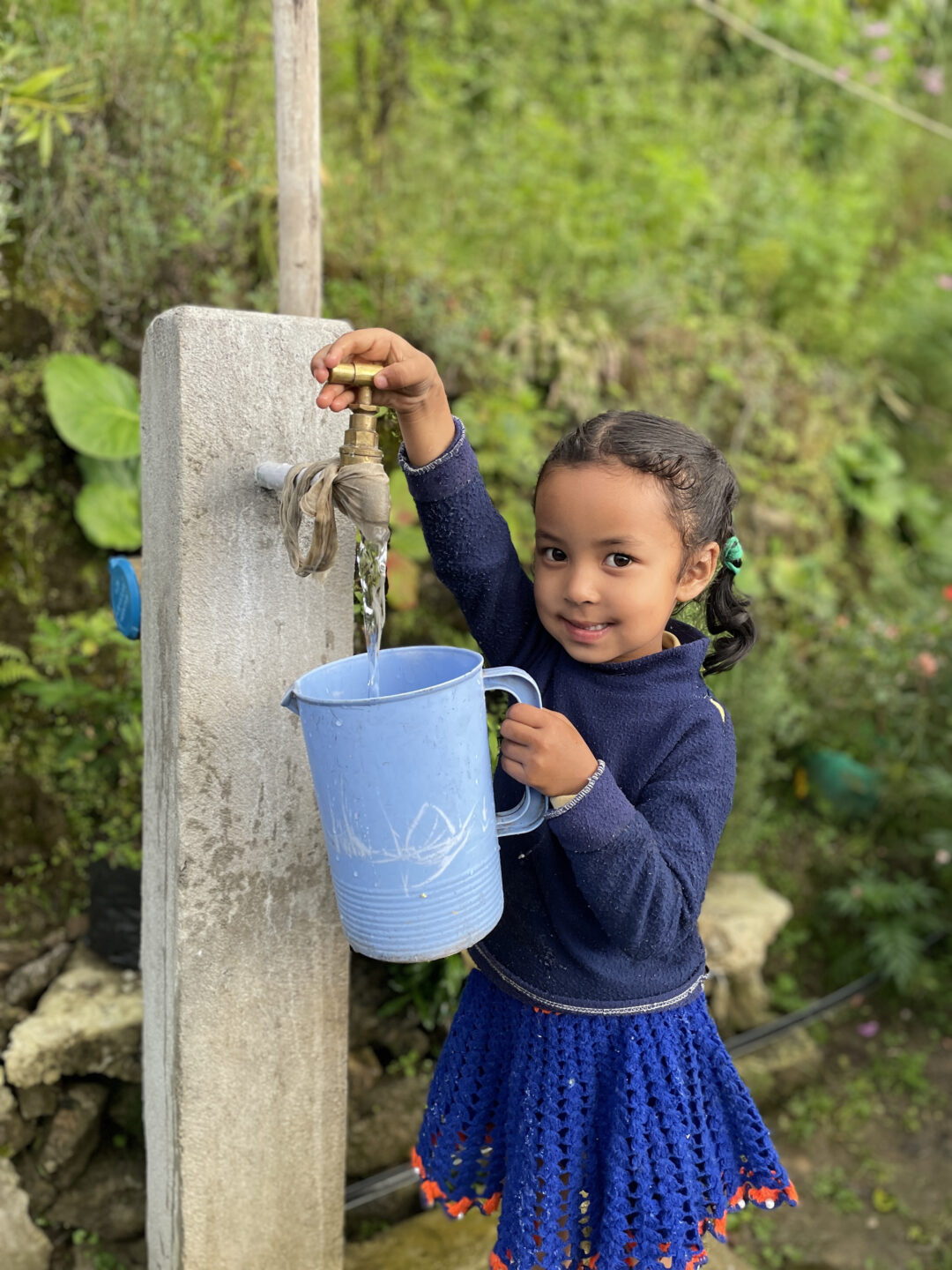 A young girl in a blue sweater and skirt smiles as she fills a blue jug with water from an outdoor tap, surrounded by lush greenery.