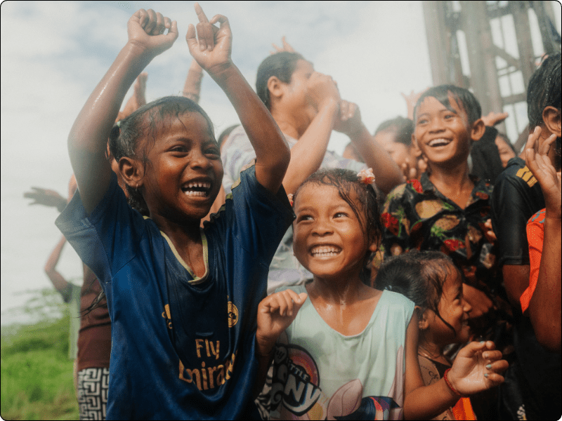 A group of joyful children, some with wet clothes and hair, smile and cheer outdoors on a cloudy day. The kids appear to be celebrating, with their hands raised and expressions of excitement and happiness. A group of joyful children, some with wet clothes and hair, smile and cheer outdoors on a cloudy day. The kids appear to be celebrating, with their hands raised and expressions of excitement and happiness.
