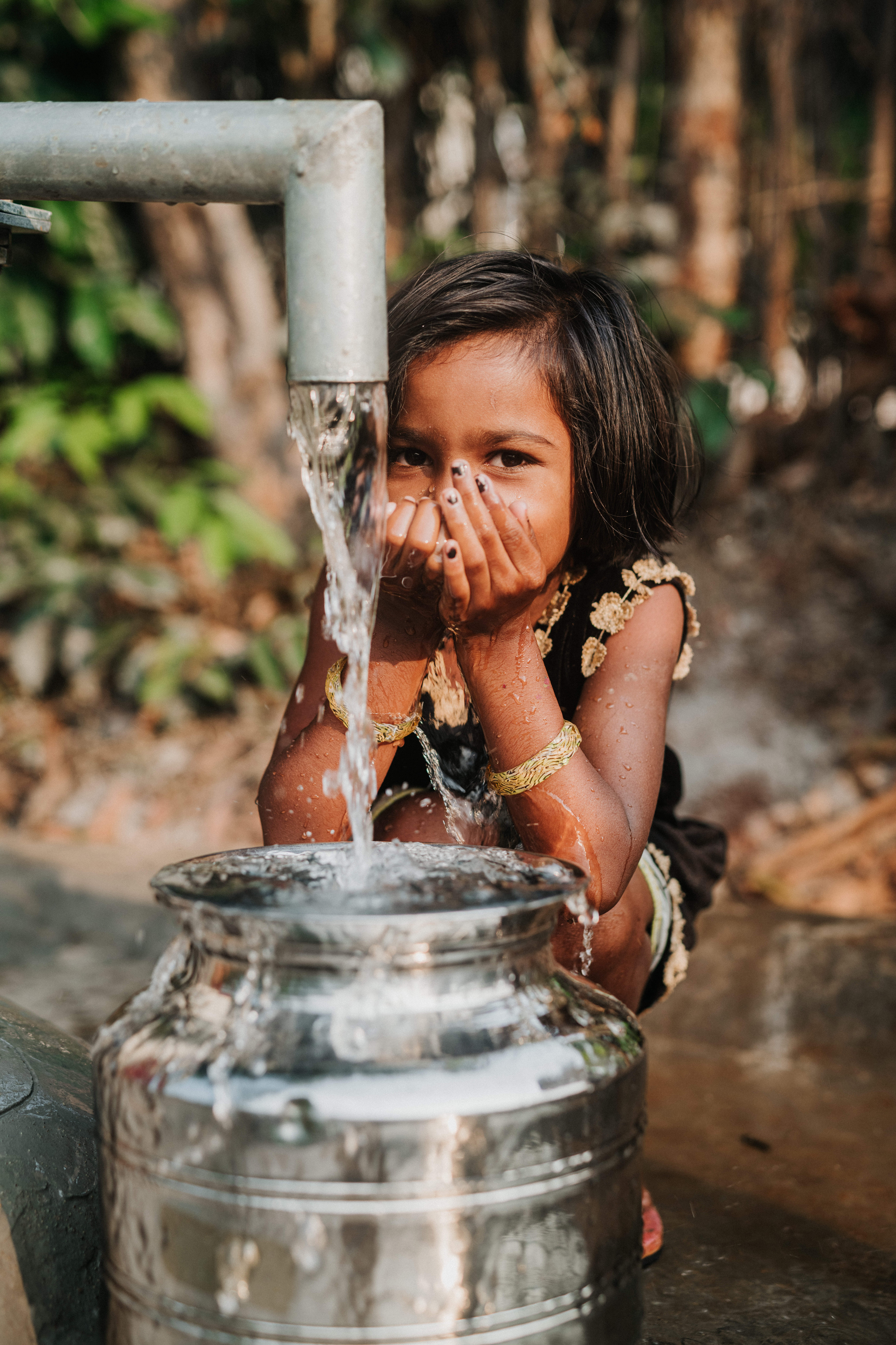 A young girl crouches by a water pump, cupping her hands to drink fresh water as it flows into a metal container. She smiles shyly, surrounded by green foliage and dappled sunlight.
